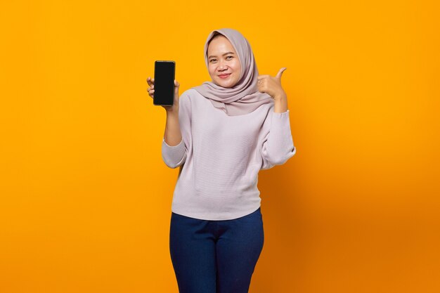 Portrait of smiling young asian woman showing smartphone blank screen and makes thumbs up sign