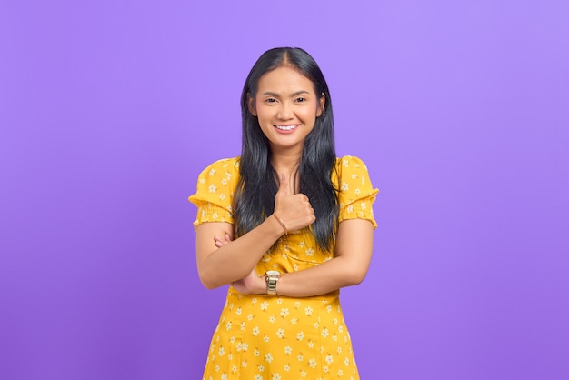 Portrait of smiling young Asian woman showing thumbs up at camera on purple background