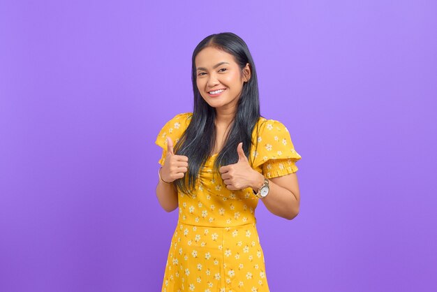 Portrait of smiling young Asian woman showing thumbs up at camera on purple background