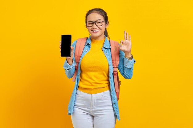 Portrait of smiling young Asian woman student in casual clothes with backpack showing blank mobile phone screen, making ok gesture isolated on yellow background