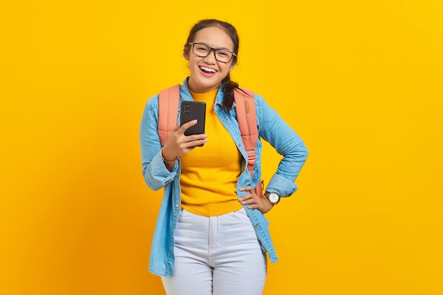 Портрет улыбающейся молодой азиатской студентки в повседневной одежде с рюкзаком, использующей мобильный телефон и смотрящей в камеру, изолированную на желтом фоне. Образование в колледже университета концепции