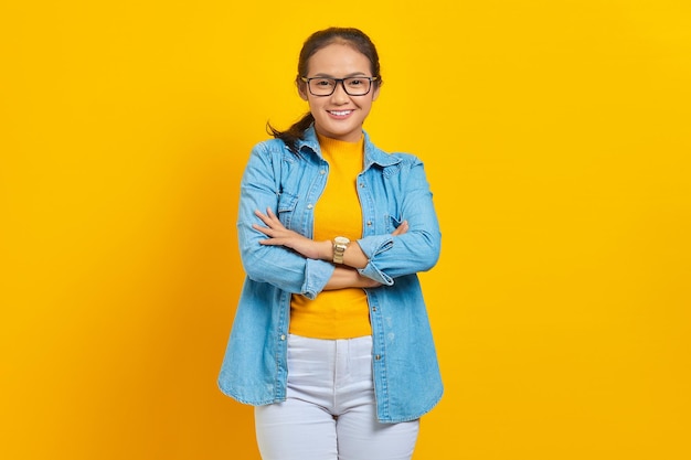 Portrait of smiling young asian woman student in denim clothes crossed arms chest and looking confident isolated on yellow background. education in college university concept