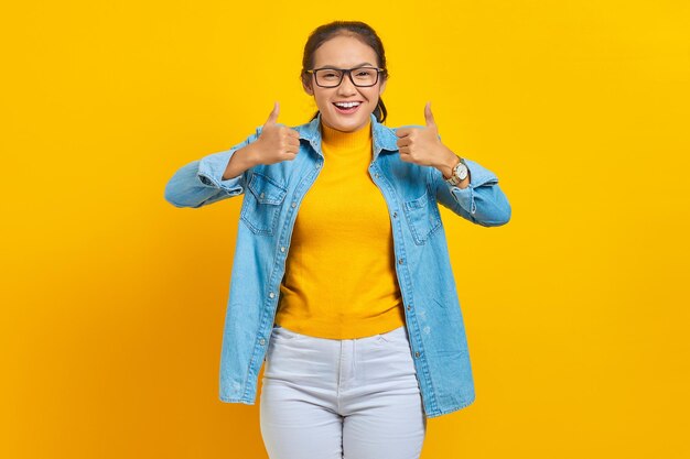 Portrait of smiling young Asian woman student in denim clothes  showing thumbs up gesture with hand isolated on yellow background. Education in college university concept