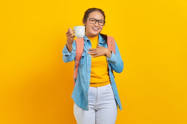 Portrait of smiling young asian woman student in denim clothes with backpack showing cup of coffee at camera isolated on yellow background