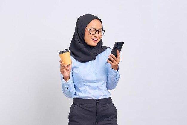 Portrait of smiling young Asian woman using a mobile phone and holding a cup of coffee isolated on white background