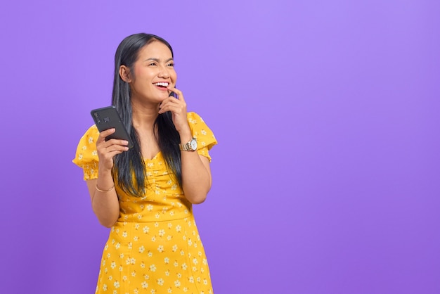 Portrait of smiling young asian woman using a mobile phone and looking away on purple background