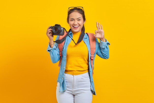 Portrait of smiling young asian woman with backpack holding professional camera and showing okay gesture isolated on yellow background