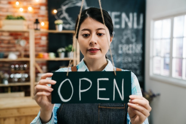 Photo portrait of smiling young barista girl in apron holding open sign board while standing at cafe bar. elegant asian coffee shop female staff turn door plate in the morning in own store small business