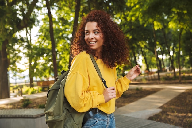 Portrait of a smiling young beautiful curly student outdoors in nature park walking with backpack.