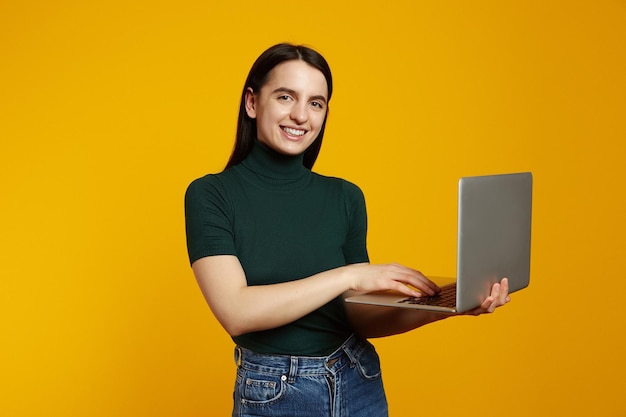 Portrait of a smiling young brunette girl holding laptop computer isolated over yellow background