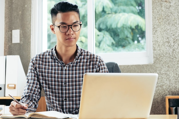 Portrait of smiling young businessman in glasses reading information in laptop screen and taking notes in planner