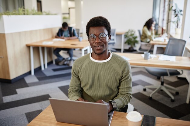 Portrait of smiling young businessman in glasses working on laptop at office desk and answering emai...