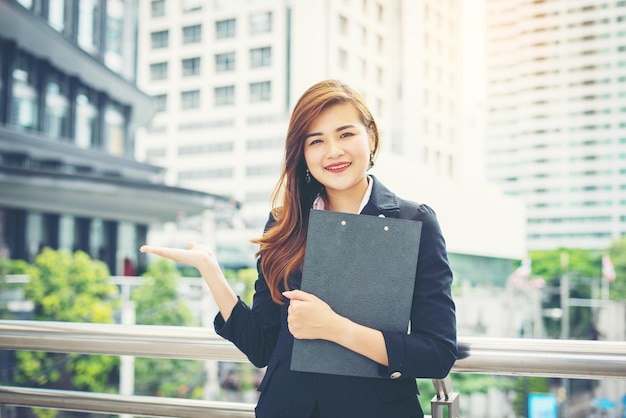 Portrait of smiling young businesswoman with folder gesturing against buildings in city