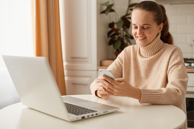 Portrait of smiling young Caucasian woman wearing beige casual style sweater with ponytail hairstyle sitting at table and holding cell phone, watching friends' photos in social networks.