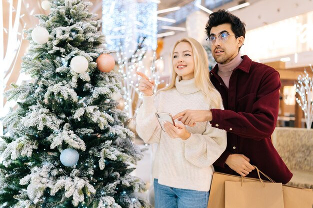 クリスマスイブにショッピング モールを祝うホールで紙袋を持って一緒に立っているスマート フォンを使用して笑顔の若いカップルの肖像画