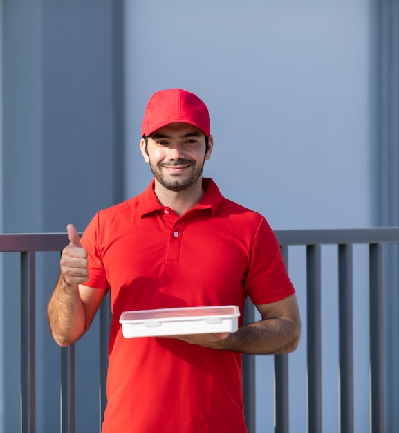 Portrait smiling young delivery man in red uniform holding a\
box in front of the house.