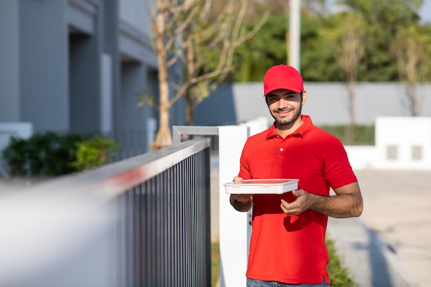 Portrait smiling young delivery man in red uniform holding a\
box in front of the house.