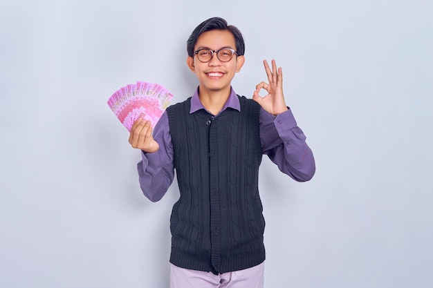 Portrait of smiling young handsome Asian businessman holding cash money in rupiah banknotes and gesturing okay sign isolated on white background People lifestyle concept