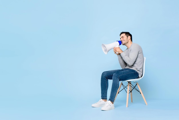 Portrait of smiling young handsome Caucasian man sitting while holding and announcing on megaphone in isolated studio blue background