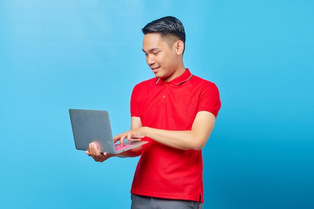 Ritratto di giovane uomo bello sorridente che tiene e utilizza un computer portatile su sfondo blu