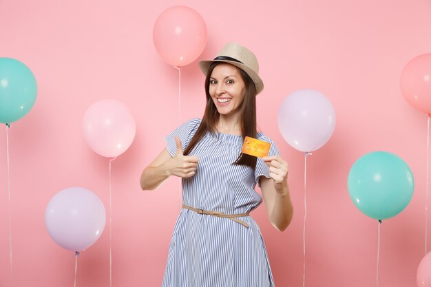 Portrait of smiling young happy woman in straw summer hat blue dress holding credit card showing thumb up on pink background with colorful air balloons. Birthday holiday party people sincere emotions.