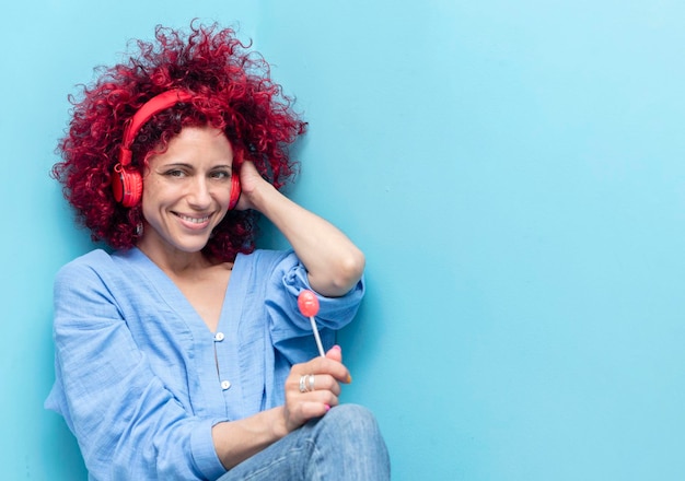 Portrait of a smiling young latin woman with a red afro hair in blue background holding a lollipop listening to music with red headphones