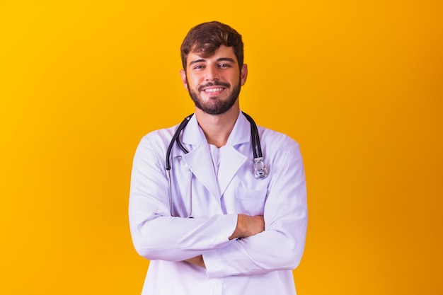 Portrait of smiling young male doctor with stethoscope around neck standing with arms crossed in white coat isolated on yellow background