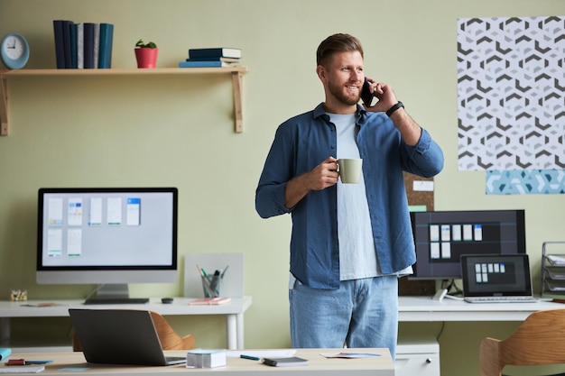 Портрет улыбающегося молодого человека, звонящего по телефону и наслаждающегося кофе во время перерыва в работе в о