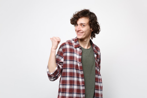 Portrait of smiling young man in casual clothes looking camera, pointing thumb aside behind his back isolated on white wall background. People sincere emotions, lifestyle concept. Mock up copy space.