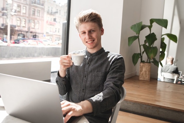 Portrait of a smiling young man dressed in a shirt sitting in a light cafe with a laptop and a cup of coffee