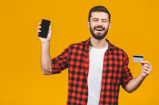 Portrait of a smiling young man holding blank screen mobile phone and showing credit card isolated over yellow wall.