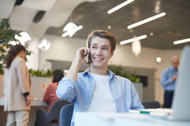 Portrait of smiling young man lspeaking by smartphone while working with laptop in office