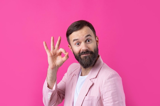 Portrait of a smiling young man in a pink jacket Man showing ok gesture while standing in studio over isolated red background