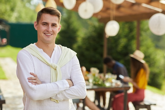 Портрет улыбающегося молодого человека, позирующего на открытом воздухе летом с друзьями и семьей, наслаждаясь ужином на террасе