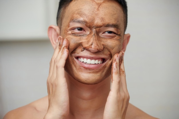 Portrait of smiling young man scrubbing face to get rid of dead\
skin cells