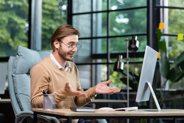 Portrait of a smiling young man sitting at the desk in the office in front of the computer and