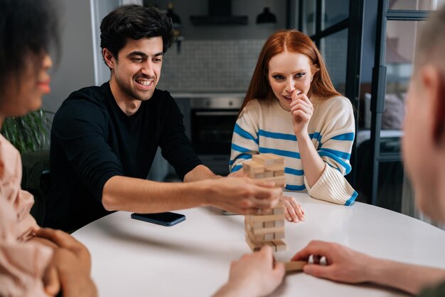 Portrait of smiling young man sitting at table in living room playing society games with multiethnic friends Joyful male pulling wooden block from wooden tower