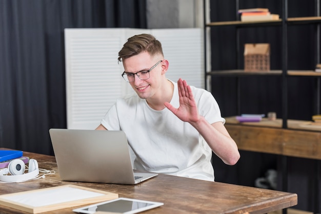 Ritratto di un giovane sorridente, agitando la mano mentre si chatta su video sul computer portatile