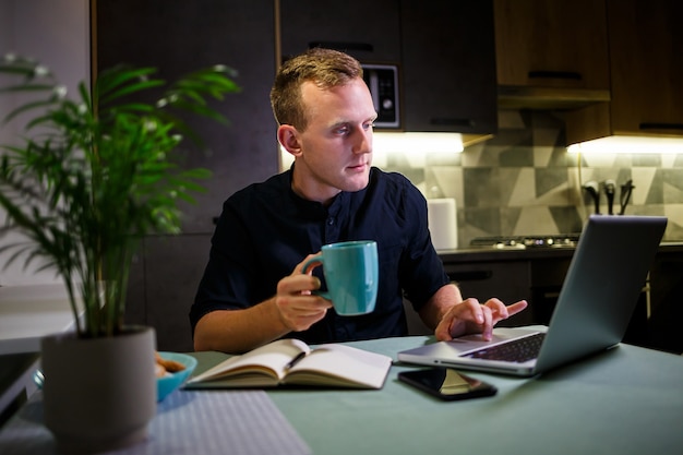 Portrait of a smiling young man working on a laptop from home and drinking coffee from a mug and is satisfied with his work. Cozy modern apartment. Home work, online business concept