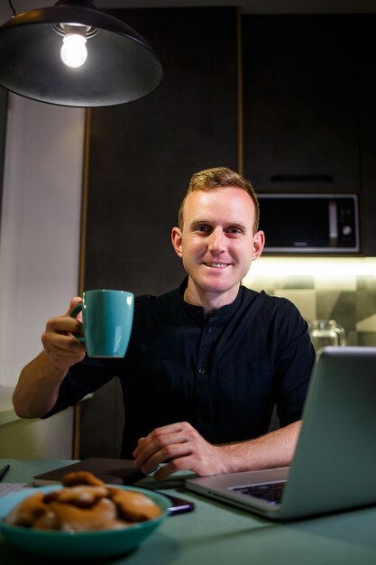 Портрет улыбающегося молодого человека, работающего на ноутбуке из дома, пьющего кофе из кружки и довольного своей работой. Уютная современная квартира. Домашняя работа, бизнес-концепция онлайн