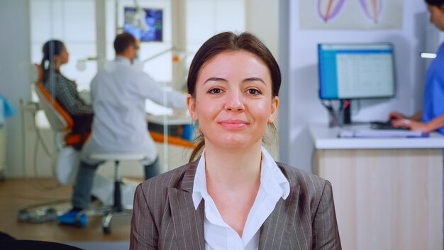 Portrait of smiling young patient looking on webcam sitting on chair in waiting room of stomatological clinic while doctor working in background. Stomatologist assistant typing on pc in dental office