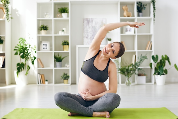 Ritratto di giovane donna incinta sorridente in abito sportivo seduto con le gambe incrociate e piegandosi da parte con il braccio alzato mentre si pratica lo yoga a casa