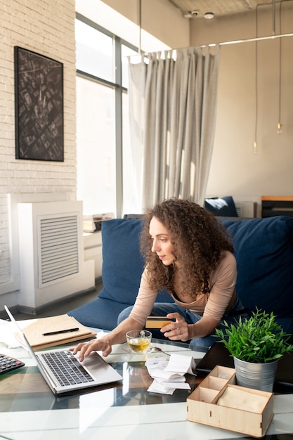 Portrait of smiling young SMM manager with curly hair sitting on comfortable sofa and working with laptop in cafe