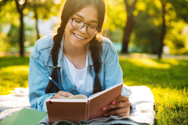 Portrait of a smiling young student girl wearing eyeglasses sitting outdoors in nature park writing notes reading book.