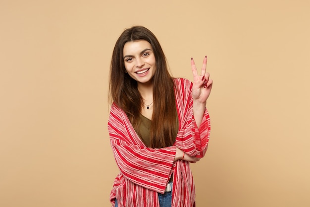 Portrait of smiling young woman in casual clothes looking camera, showing victory sign isolated on pastel beige background in studio. People sincere emotions, lifestyle concept. Mock up copy space.