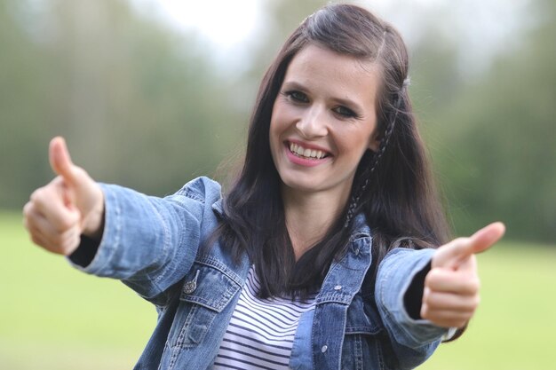 Foto ritratto di una giovane donna sorridente che fa un gesto con i pollici contro gli alberi