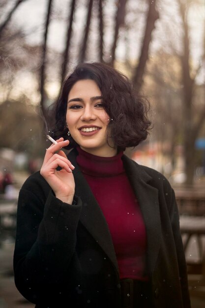 Foto ritratto di una giovane donna sorridente che tiene una sigaretta in piedi contro gli alberi