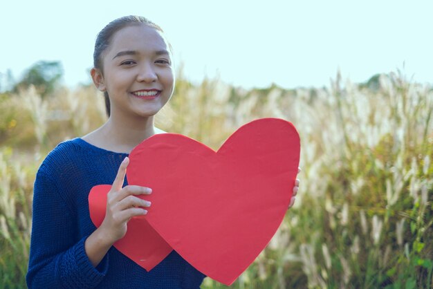 Photo portrait of a smiling young woman holding heart shape outdoors