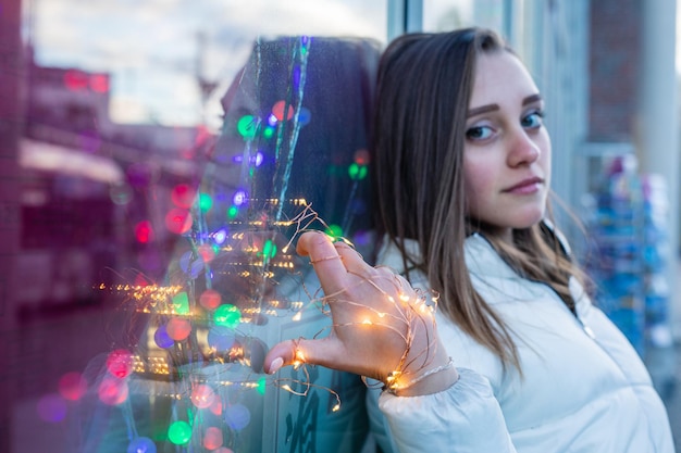 Portrait of smiling young woman holding illuminated lighting equipment while leaning on glass window