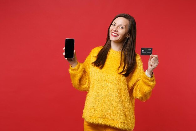 빈 화면이 있는 휴대폰을 들고 웃고 있는 젊은 여성의 초상화, 밝은 빨간색 벽 배경에 격리된 신용 은행 카드. 사람들은 진심 어린 감정, 라이프 스타일 개념입니다. 복사 공간을 비웃습니다.